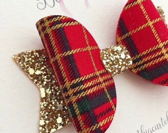 Puede incluir: Un lazo rojo a cuadros con detalles de purpurina dorada. El lazo presenta un patrón de tartán clásico en rojo, verde y negro, con hilos dorados. Debajo del lazo se ve una forma de estrella de purpurina dorada.
