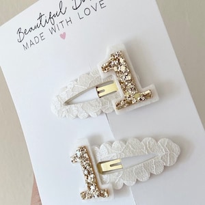 May include: Two white hair clips with a gold glitter number one on each. The clips are decorated with a white lace design. The text "Beautiful Bows Made With Love" is printed on a white card behind the clips.