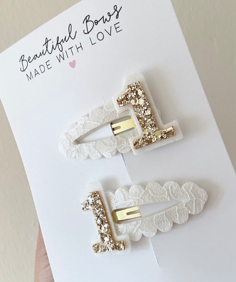 May include: Two white hair clips with a gold glitter number one on each. The clips are decorated with a white lace design. The text "Beautiful Bows Made With Love" is printed on a white card behind the clips.