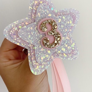 May include: A layered star-shaped decoration with glitter and a pink ribbon. The star has a light purple and white colour scheme with a gold glitter number 3 in the centre. This item is likely for a birthday celebration.