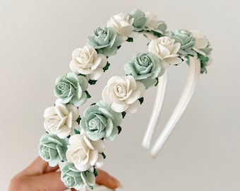 May include: A white headband with two rows of green and white paper flowers. The flowers are arranged in a circle around the headband.