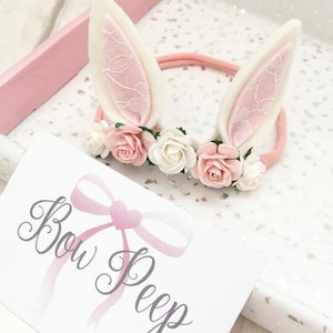May include: A pink headband with white bunny ears and a floral crown of pink and white flowers. The headband is on a white surface with a pink and white card that says "Bow Peep".
