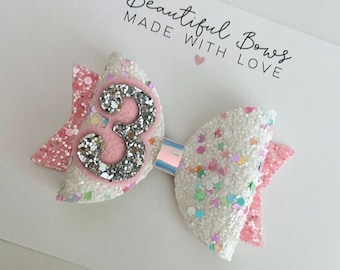 May include: A white and pink glitter bow with a silver number 3 in the centre. The bow has a rainbow iridescent ribbon in the centre. The bow is on a white background with the text "Beautiful Bows Made With Love".