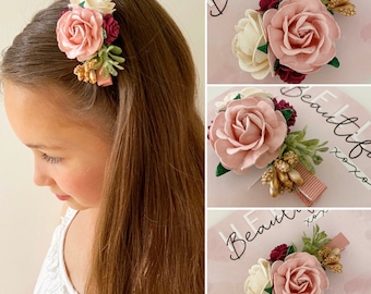 Puede incluir: Una pinza para el pelo con flores rosas y blancas y detalles dorados. La pinza está sujeta al cabello castaño de una niña. El texto "Hello Beautiful xoxo" es visible en el fondo.