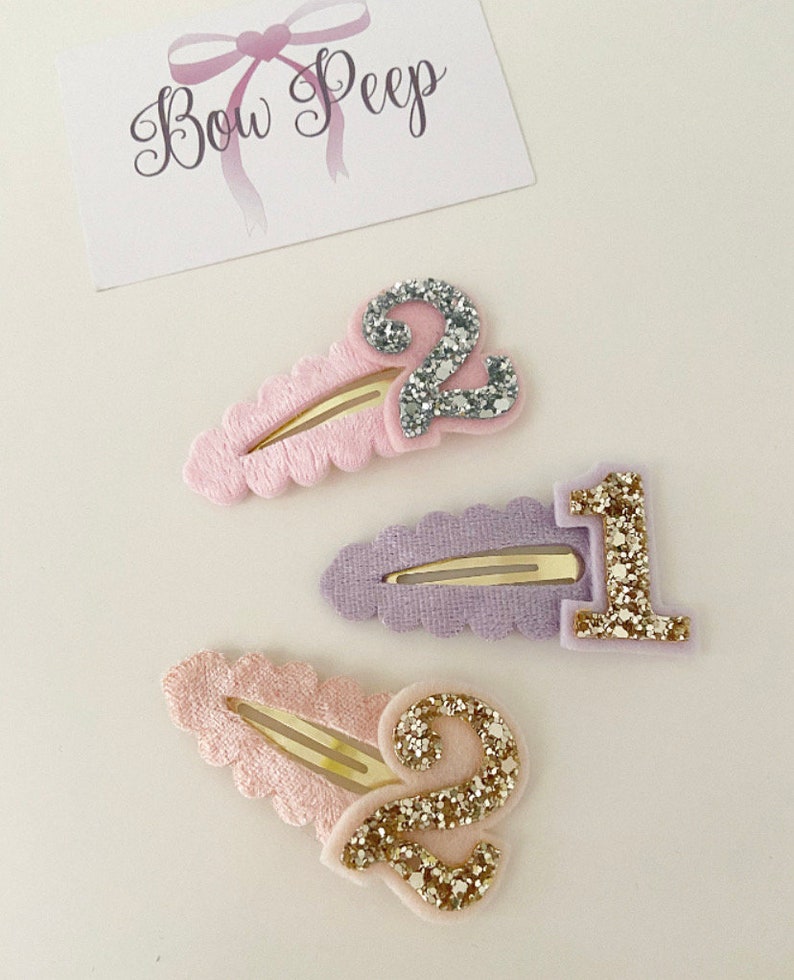 May include: Three glitter hair clips with numbers 1 and 2. The clips are pink, purple, and light pink with gold and silver glitter.