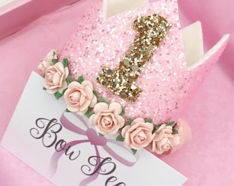 Puede incluir: Corona de purpurina rosa con un número uno de purpurina dorada y una corona de flores. La corona dice "Bow Peep" en una etiqueta blanca.