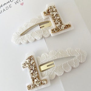May include: Two white hair clips with gold glitter number one embellishments. The clips have a scalloped edge and are made with a white lace fabric.
