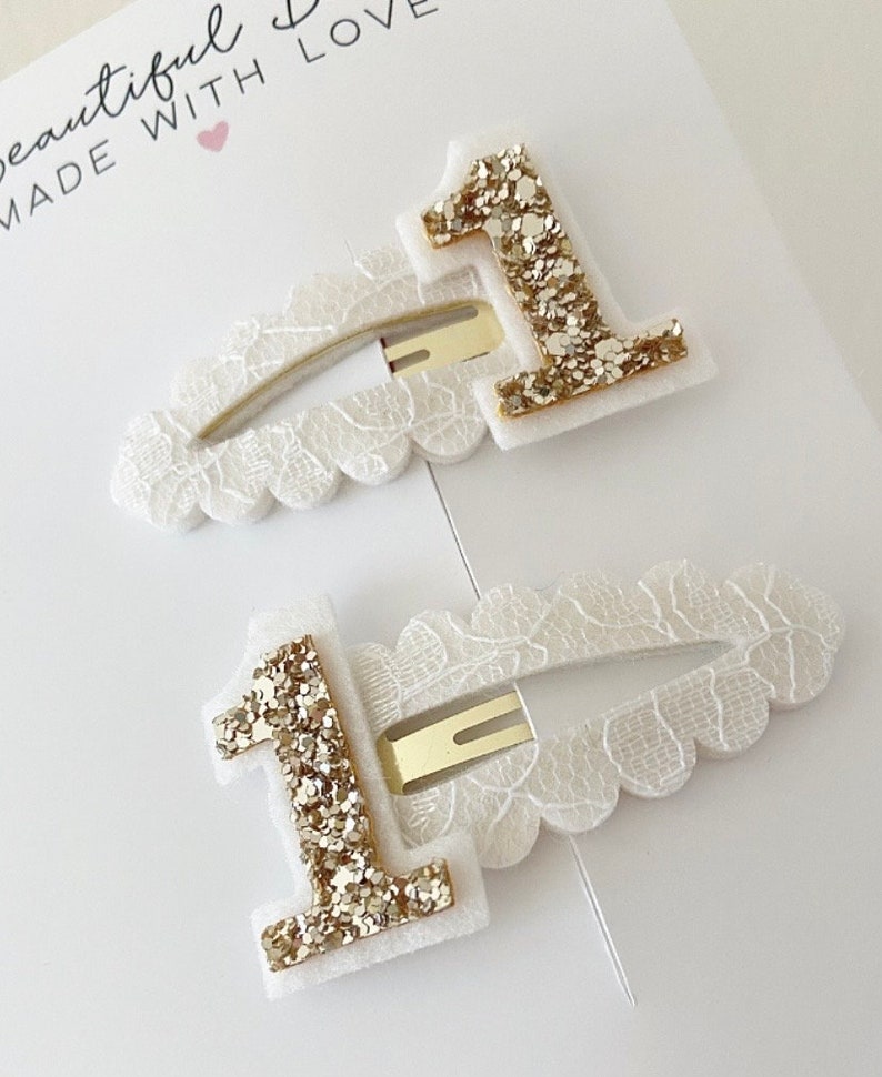 May include: Two white hair clips with gold glitter number one embellishments. The clips have a scalloped edge and are made with a white lace fabric.