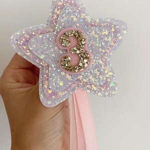 May include: A star-shaped wand with glitter in white, pink, and lavender, displaying the number "3" in gold glitter. It has a pink ribbon attached. This item is suitable for a birthday celebration.