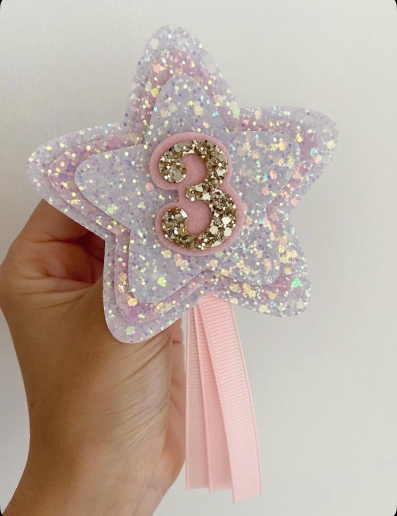 May include: A star-shaped wand with glitter in white, pink, and lavender, displaying the number "3" in gold glitter. It has a pink ribbon attached. This item is suitable for a birthday celebration.