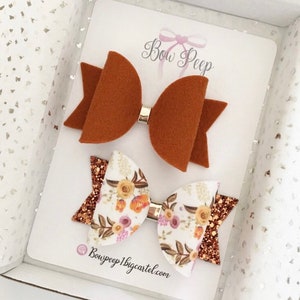 May include: Two hair bows presented in a white box. One is a solid brown colour, the other features a floral pattern on white with glitter accents. Both are attached to silver clips. The text "Bow Peep" is visible.