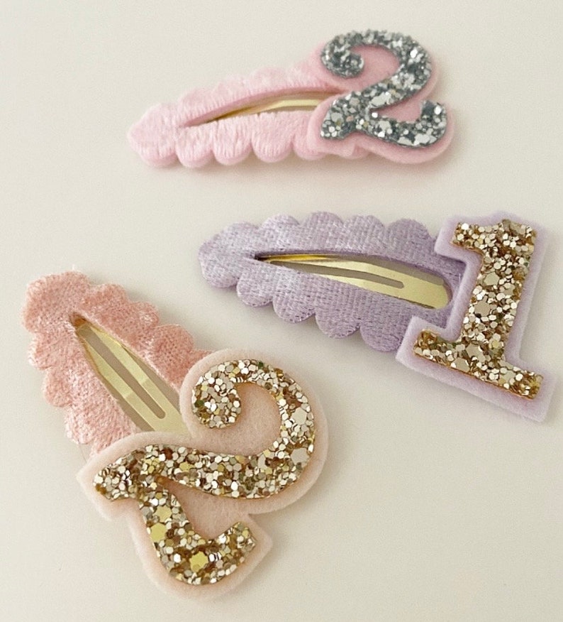 May include: Three hair clips with velvet fabric and glitter numbers. The clips are pink, purple, and pink with gold glitter numbers 2, 1, and 2.
