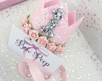 Puede incluir: Corona de fieltro rosa con un número uno de purpurina plateada y un borde floral. La corona está unida a una diadema elástica rosa. La corona está en una caja con una etiqueta que dice "Bow Peep".