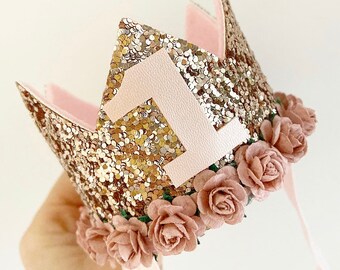 Puede incluir: Una corona dorada con purpurina con un número 1 rosa, ribete rosa y pequeñas flores de papel rosas. La corona tiene cintas rosas adjuntas para atar. Esta corona festiva es perfecta para una primera celebración de cumpleaños.