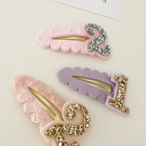 May include: Three hair clips with numbers 1 and 2 on them. The clips are pink, purple, and light pink with gold glitter numbers.