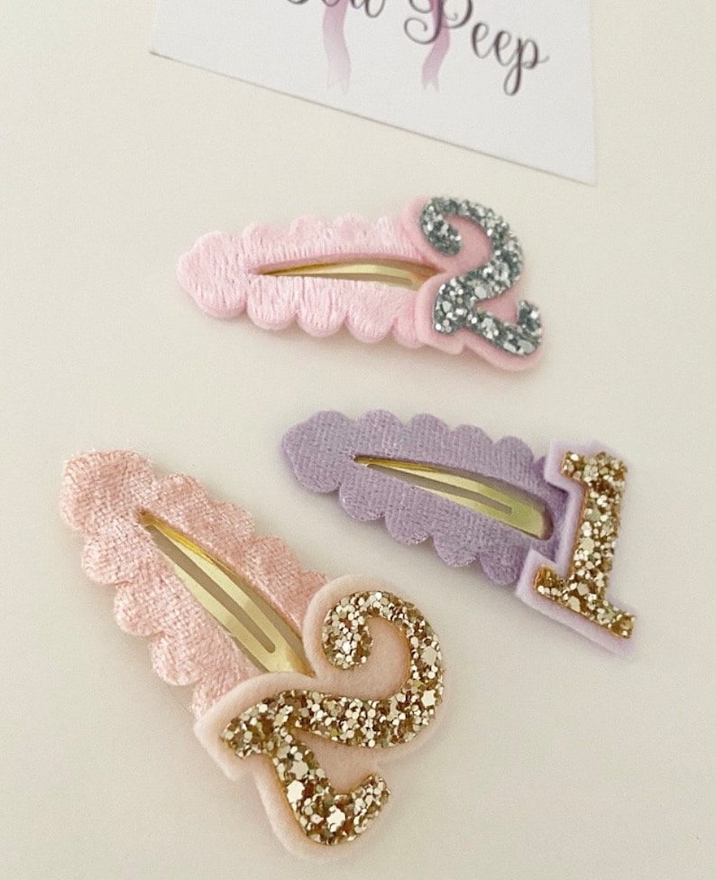 May include: Three hair clips with numbers 1 and 2 on them. The clips are pink, purple, and light pink with gold glitter numbers.