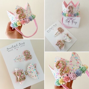 Puede incluir: Un conjunto de tres accesorios para el cabello de purpurina rosa y blanca con adornos de purpurina dorada número 2. El conjunto incluye una diadema, un lazo y una pinza para el cabello. La diadema es una corona rosa con una base de purpurina blanca y un número 2 de purpurina dorada. El lazo es un lazo rosa con una base de purpurina blanca y un número 2 de purpurina dorada. La pinza para el cabello es una base de purpurina blanca con un número 2 de purpurina dorada. El conjunto está etiquetado como "Beautiful Bows Made With Love".