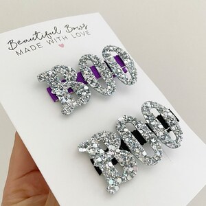 May include: Two glittery silver hair clips spelling "BOO" on a white card. The letter "B" is solid, while the "OO" are open ovals. The clips have purple and black accents. The card reads "Beautiful Bows MADE WITH LOVE".