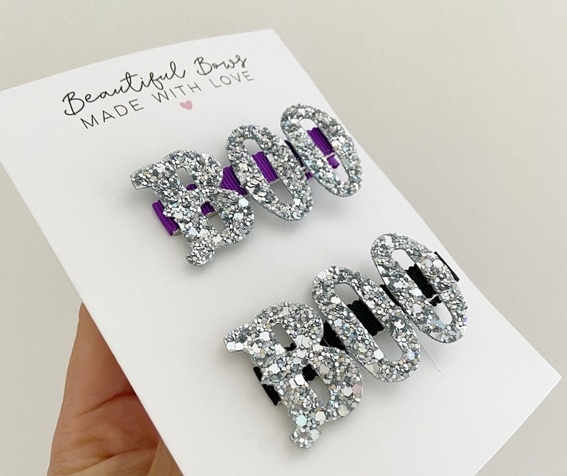 May include: Two glittery silver hair clips spelling "BOO" on a white card. The letter "B" is solid, while the "OO" are open ovals. The clips have purple and black accents. The card reads "Beautiful Bows MADE WITH LOVE".