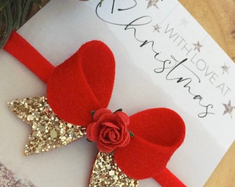 May include: Red velvet bow with gold glitter accents and a red rose in the centre. The bow is attached to a red headband. The background is a white card with the text "With Love at Christmas".