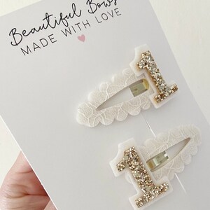 May include: Two white lace hair clips with gold glitter number one embellishments. The clips are packaged on a white card with the text "Beautiful Bows Made With Love" and a pink heart.