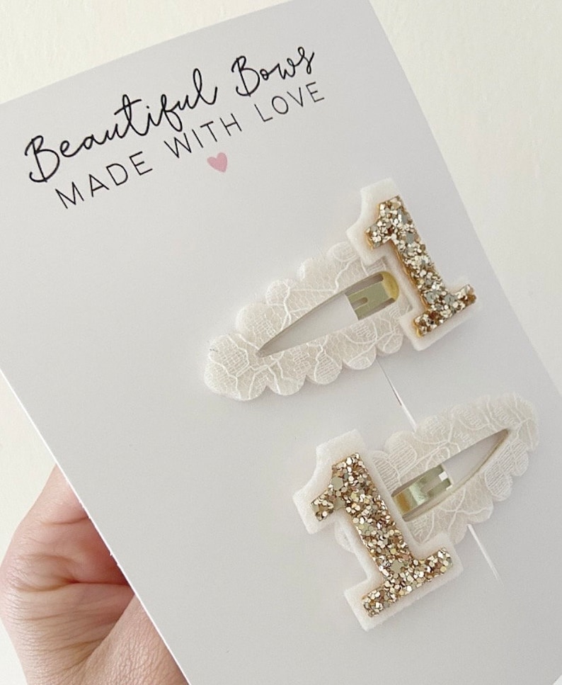 May include: Two white lace hair clips with gold glitter number one embellishments. The clips are packaged on a white card with the text "Beautiful Bows Made With Love" and a pink heart.