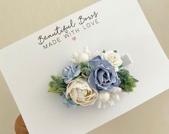 Puede incluir: Pinza para el pelo floral con flores de tela azules, crema y blancas, hojas verdes y detalles blancos. La pinza está sobre una tarjeta blanca con el texto "Beautiful Bows MADE WITH LOVE".