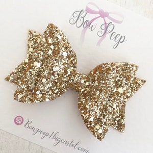May include: Gold glitter hair bow with a sparkly finish. The bow is attached to a clip.