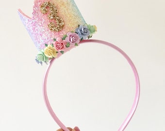 Puede incluir: Una diadema rosa con una corona de purpurina arcoíris y un número 3 dorado. La corona está decorada con flores de colores pastel.