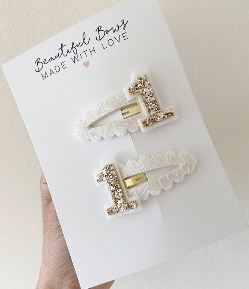 May include: Two white hair clips with gold glitter number one embellishments. The clips are displayed on a white card with the text "Beautiful Bows Made With Love".