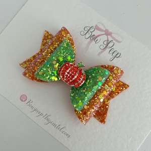 May include: A layered hair bow featuring orange and green glitter fabric, centred with a small red rhinestone pumpkin embellishment. The bow is attached to a white display card printed with the text Bow Peep and Bowpeep1.bigcartel.com.