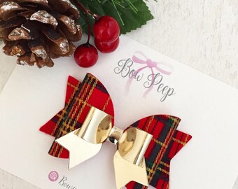 Puede incluir: Un lazo para el pelo a cuadros rojos, verdes y dorados, con un centro dorado brillante. El lazo está sobre una tarjeta blanca con el texto "Bow Peep" y "Bowpeep1bigcartel.com". Una piña, bayas y vegetación en el fondo.