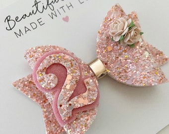 May include: A pink glitter hair bow with a pink felt number "2" in the centre. The bow has a small white flower with green leaves on top. The bow is attached to a gold clip.