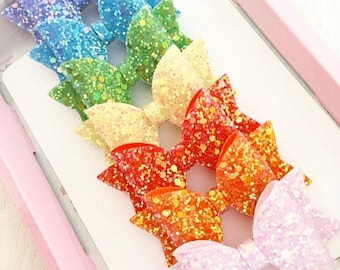 May include: A set of seven rainbow glitter hair bows. The bows are arranged in a rainbow order from purple to pink. Each bow is made of a different colour glitter.