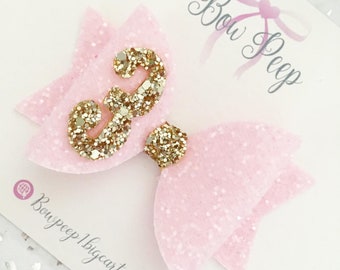 May include: Pink glitter bow with a gold glitter number 3 in the centre. The bow is on a white background with the text "Bow Peep" written in cursive.