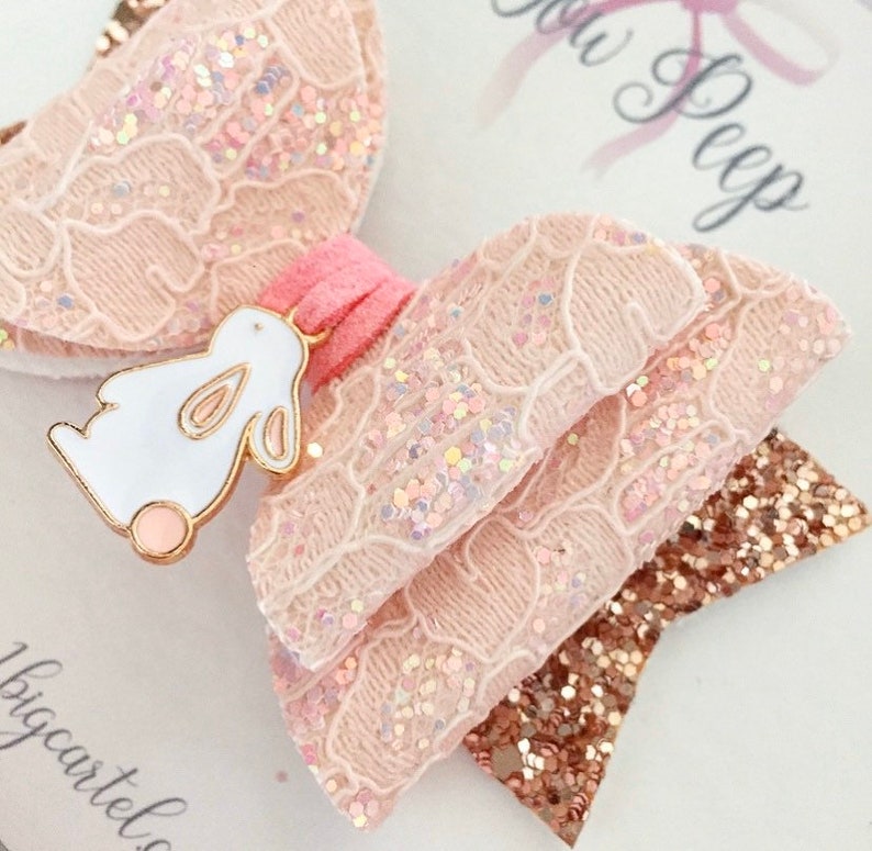 Puede incluir: Un lazo de brillo rosa y dorado con un amuleto de conejo blanco. El lazo est&aacute; hecho de encaje y tiene una cinta rosa en el centro.
