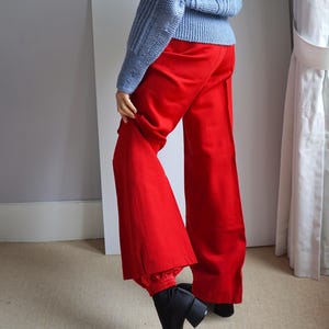 May include: A pale blue knitted jumper with a ribbed texture is worn with vibrant red wide-leg trousers. The trousers have a high waist and a flared design. Black ankle boots complete the outfit, creating a bold colour contrast.