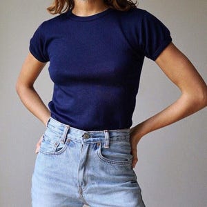 May include: A dark navy blue short-sleeved t-shirt with a crew neckline and a fitted silhouette, paired with light-wash blue denim jeans featuring a high-waisted cut, a metal button closure, and visible front pockets with copper-coloured rivets.