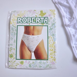 Puede incluir: Un slip de encaje blanco con un patrón floral geométrico dentro de un paquete de plástico transparente. El empaque presenta la marca ROBERTA en letras verdes, un borde floral y una fotografía del slip puesto. El número 34332 está impreso.