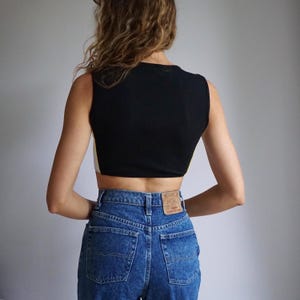 May include: A black sleeveless crop top with a white trim and a high neckline. The top is paired with blue high-waisted jeans.