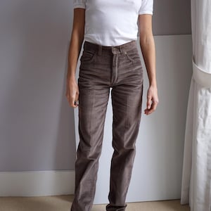 Pode incluir: Uma pessoa vestindo uma camiseta branca de manga curta e calças de veludo cotelê marrons. As calças têm um design de perna reta e um zíper frontal. A roupa é completada com sapatos pretos. O fundo é uma parede cinza lisa.