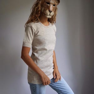 May include: A woman wearing a light grey short-sleeved t-shirt with a round neckline and blue jeans. She is wearing a brown animal mask with a white snout.