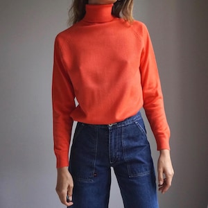 Op de afbeelding: Een feloranje coltrui gecombineerd met een donkerblauwe spijkerbroek. De trui heeft lange mouwen en een geribbelde kraag. De jeans heeft een hoge taille en grote voorzakken. Een eenvoudige, klassieke outfit.