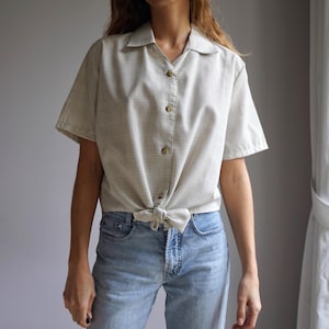 Peut inclure: Chemise beige à manches courtes avec un motif à carreaux discrets, boutonnée et nouée à la taille. Elle est portée avec un jean bleu clair. Le col de la chemise est classique.