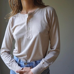 Könnte beinhalten: Beiges Langarm-Henley-Shirt mit einer Knopfleiste mit drei Knöpfen. Das Shirt hat einen Rundhalsausschnitt und ist aus einem weichen, leichten Stoff gefertigt. Die Ärmel sind lang und eng anliegend, getragen mit blauen Jeans.