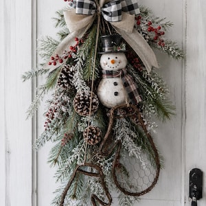 May include: A holiday swag featuring a snowman wearing a black top hat and plaid scarf, accented with pine branches, frosted pinecones, red berries, and a large burlap bow with a black and white buffalo check pattern, plus two wire-framed mittens.