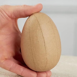May include: A light brown, egg-shaped craft item with vertical lines, held in a hand. The egg has a smooth surface and is likely made of paper or cardboard. The background is a neutral color.