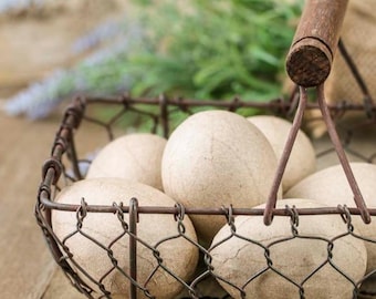 May include: A rectangular wire basket with a wooden handle containing several smooth, light beige, oval-shaped objects. The basket is constructed from dark brown metal wire in a hexagonal mesh pattern, holding the objects securely within its frame.