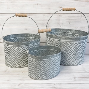 May include: Set of three galvanized metal buckets with chevron pattern and wooden handles. The buckets are different sizes, with a silver finish and a rustic aesthetic. The handles are made of metal and wood.