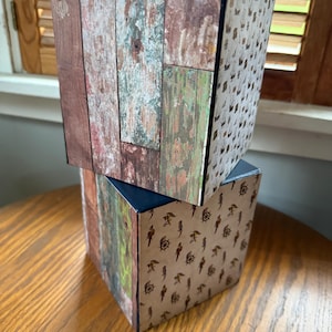 May include: Two decorative tissue box covers. One features a weathered wood design in shades of brown, green, and gray. The other has a tan background with a pattern of ships' wheels and parrots. The boxes are stacked.